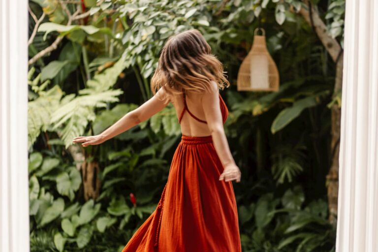 Woman in a flowing red backless dress twirls with her arms outstretched in a lush tropical garden filled with large green leaves. The movement of her hair and skirt creates a sense of freedom and connection with nature. Showing feminine embodiment