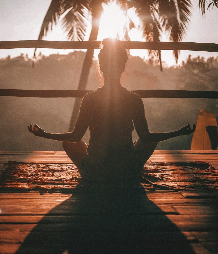 Silhouette of a person meditating cross legged on a wooden deck at sunrise with palm trees in the background. Warm golden light surrounds the figure, emphasizing a peaceful and mindful moment.