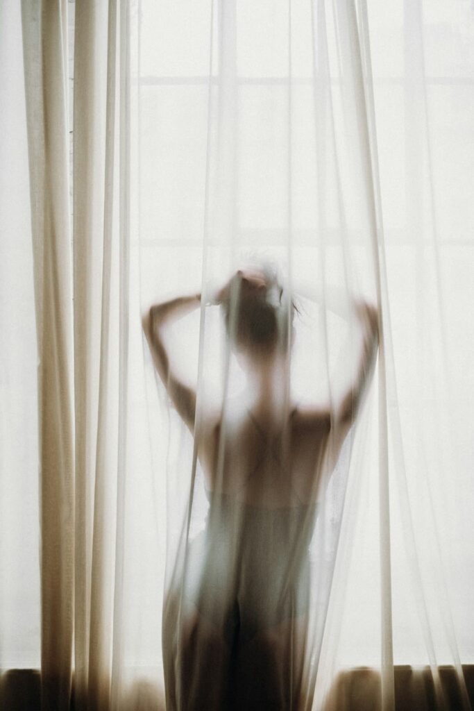 Soft blurred silhouette of a woman standing behind sheer white curtains with her arms raised as natural light filters through a window. The translucent fabric and muted tones create a quiet intimate atmosphere.