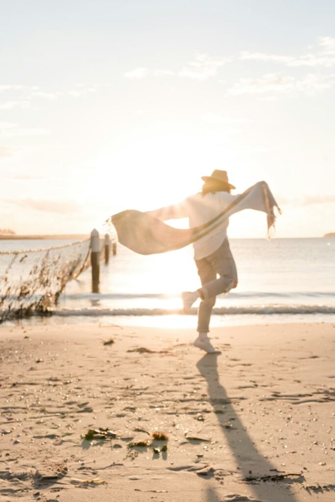 Person wearing a hat and light clothing runs along a sandy beach at sunset with a scarf flowing behind them. Long shadows stretch across the sand, capturing a carefree and joyful coastal moment.