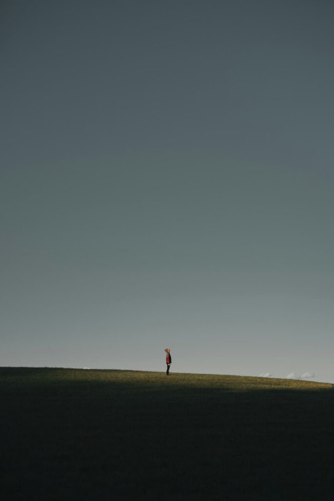 Small figure standing alone on a grassy hill beneath a vast clear blue sky. The wide open space highlights solitude and reflection in a minimalist landscape.