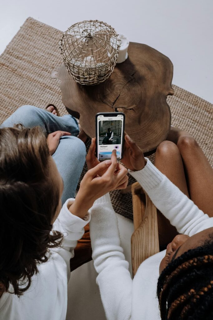 Two women sit around a wooden coffee table while one of them holds a smartphone showing a dating app profile for someone named Michael. The shot is taken from above, highlighting their casual interaction.