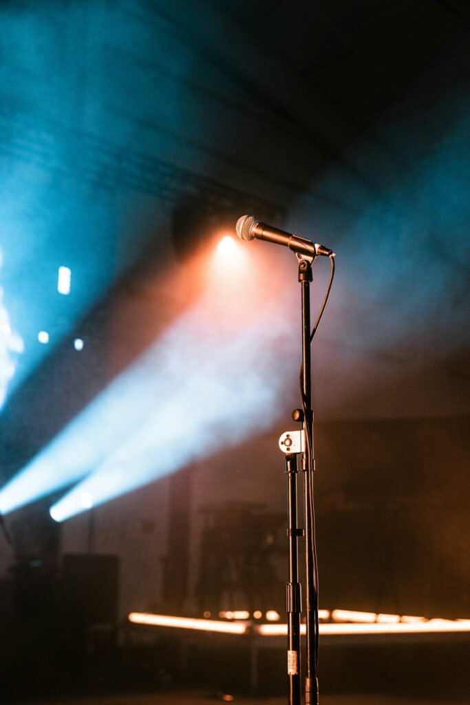 A microphone on a stand is lit by dramatic stage lighting in hues of blue and orange, with fog or smoke enhancing the concert-like atmosphere.