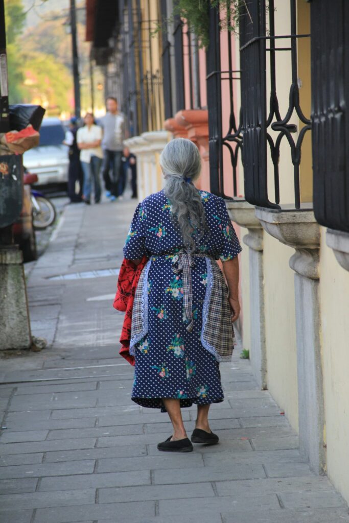 An elderly woman with gray hair tied back walks down a sidewalk in a colorful town. She wears a blue dress with floral patterns and carries a red cloth.