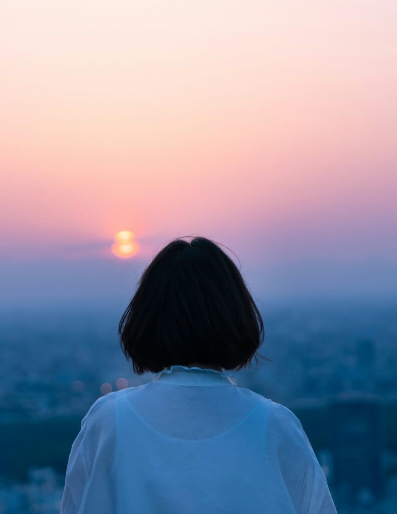 A woman with short black hair, wearing a light white blouse, stands with her back to the camera while watching the sun set over a hazy cityscape.