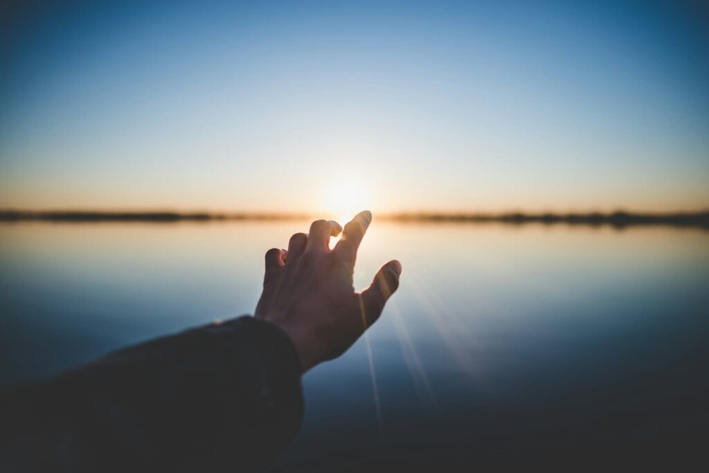 A hand extends out toward the rising or setting sun over a still lake, creating a hopeful and serene moment with a soft glow across the water.