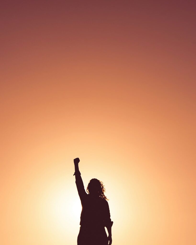 A person stands in silhouette with one arm raised triumphantly against a warm orange sunset, capturing a moment of empowerment or victory.
