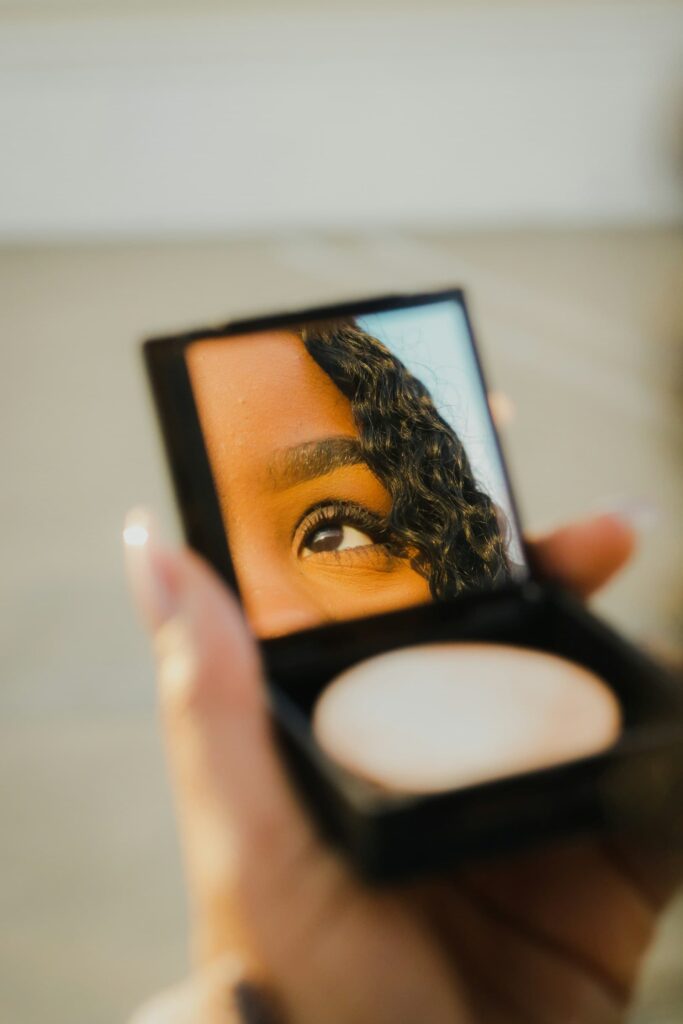 A close-up of a compact mirror reflects a woman’s eye and part of her curly hair, emphasizing her gaze and the act of self-reflection.