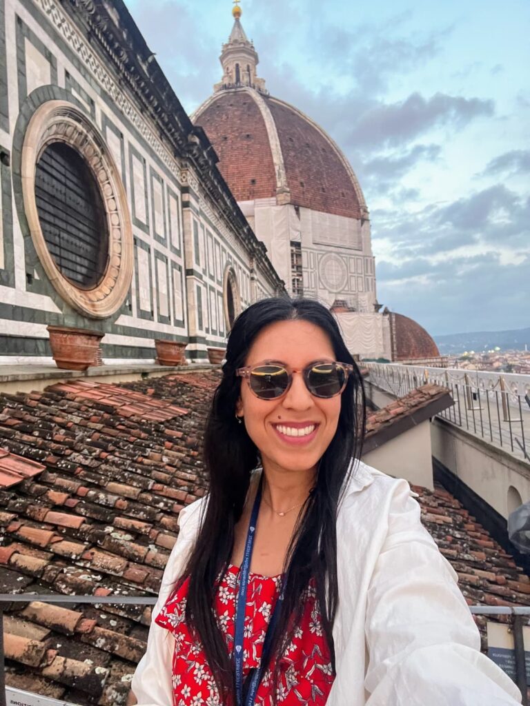 Smiling woman in sunglasses and a white jacket taking a selfie in front of the Florence Cathedral's iconic red-tiled dome at sunset. On a solo trip before dating after divorce