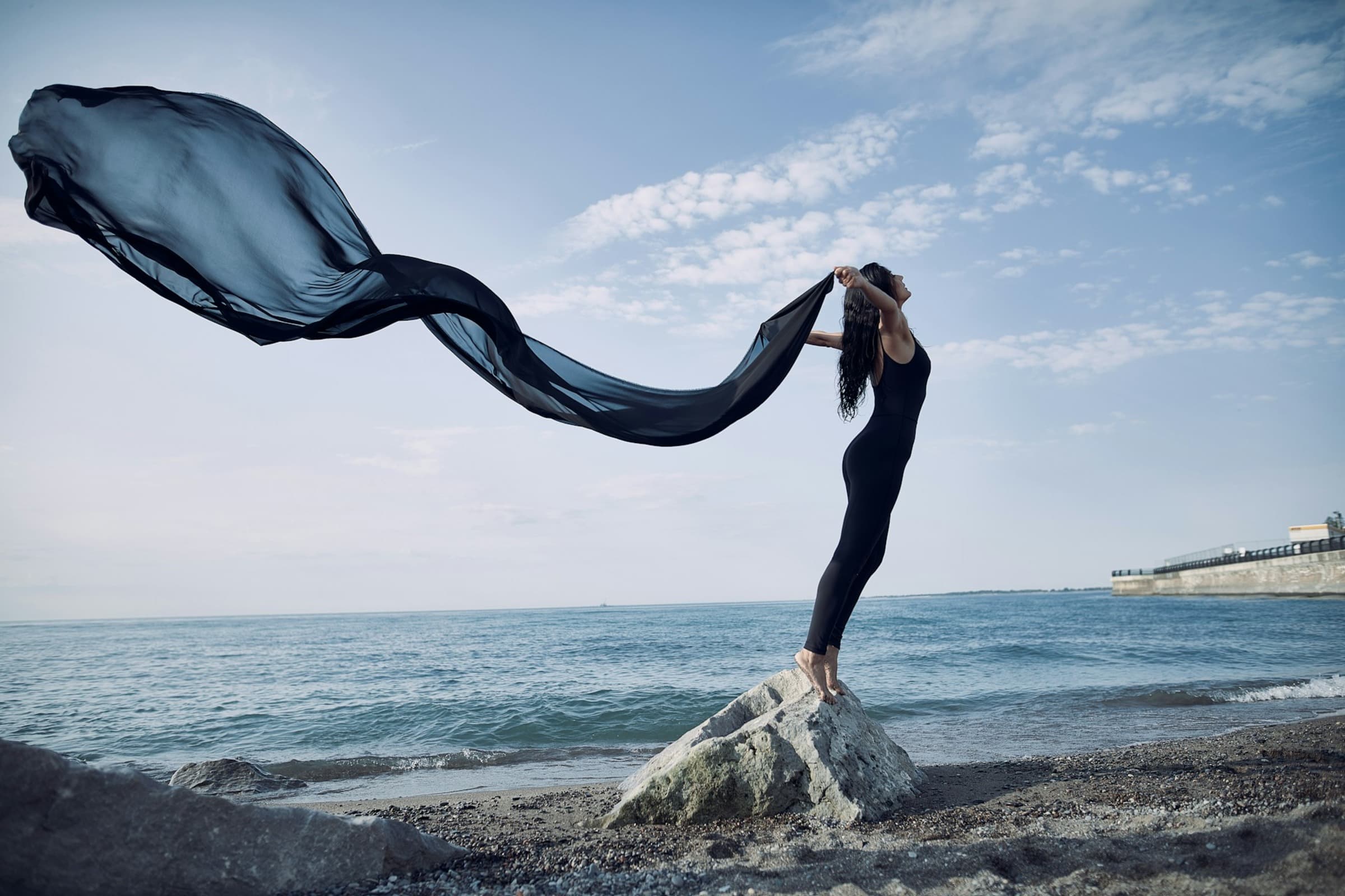 Woman standing barefoot on a rock at the beach, holding a long flowing black fabric that trails in the wind, with the ocean and sky in the background.